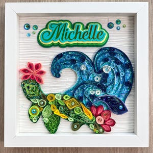 Puede incluir: Una pieza de arte en filigrana de papel enmarcada con el nombre Michelle en letra turquesa y verde. Debajo del nombre, una tortuga marina verde y amarilla con flores rosas nada frente a una gran ola azul brillante hecha de papel.
