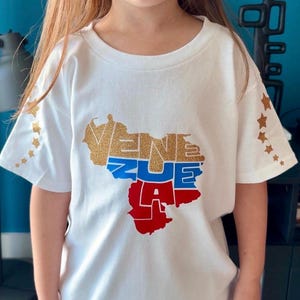 Puede incluir: Una camiseta blanca de manga corta con un mapa de Venezuela relleno con el texto VENEZUE LA en purpurina dorada, azul y rojo. Las mangas están decoradas con una fila vertical de pequeñas estrellas de purpurina dorada a cada lado.