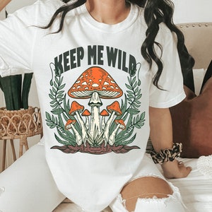 May include: White t-shirt with the text "Keep Me Wild" printed in black. The shirt features a graphic of a cluster of mushrooms with orange caps and white stems.