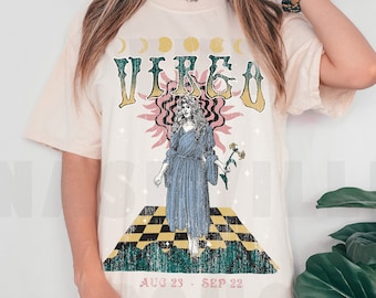 Puede incluir: Una camiseta de color crema con un gráfico de estilo vintage del signo del zodíaco Virgo. El gráfico presenta a una mujer con el pelo largo, un vestido azul y una flor en la mano. El texto "VIRGO" está en una fuente retro en negrita, y las fechas "AUG 23 - SEP 22" están impresas debajo del gráfico.