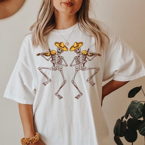 May include: White oversized t-shirt with a graphic of two skeletons wearing sombreros and playing violins.