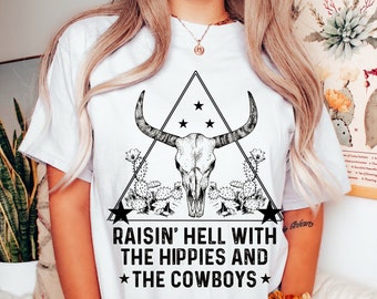 Puede incluir: Camiseta blanca con un gráfico en blanco y negro de una calavera con cuernos en un triángulo con estrellas y cactus. El texto "Raisin' Hell With The Hippies And *The Cowboys*" está impreso debajo del gráfico.