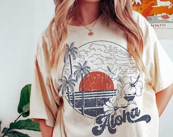 Peut inclure: T-shirt crème avec un motif circulaire représentant un coucher de soleil, des palmiers et des fleurs d'hibiscus. Le mot "Aloha" est écrit dans une police stylisée. Le t-shirt a des manches courtes et une coupe décontractée.