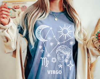 Puede incluir: Una camiseta azul de gran tamaño con un diseño gráfico blanco que presenta el signo del zodíaco Virgo, una luna creciente, un sol y estrellas. El texto "VIRGO" está impreso debajo del diseño.