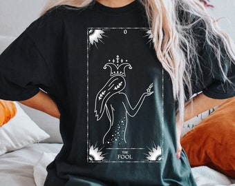 May include: Black t-shirt with a white graphic of the tarot card "The Fool". The card features a stylized image of a person with a crown and a jester's hat, holding a flower.