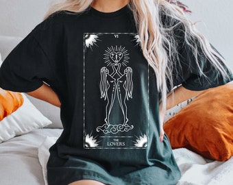 May include: Black t-shirt with a white graphic of the Lovers tarot card. The card features two figures standing on clouds with a heart above their heads. The text "THE LOVERS" is below the figures.