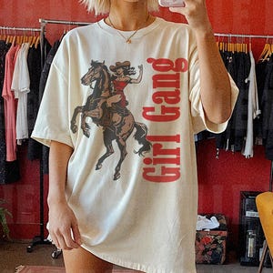 May include: A woman wearing a white t-shirt with a red graphic of a woman riding a horse and the text "Girl Gang". She is wearing pink cowboy boots.