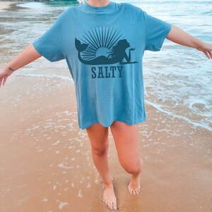 May include: A woman wearing a blue t-shirt with a mermaid and sun graphic and the word "SALTY" printed on it. She is standing on a sandy beach with the ocean in the background.