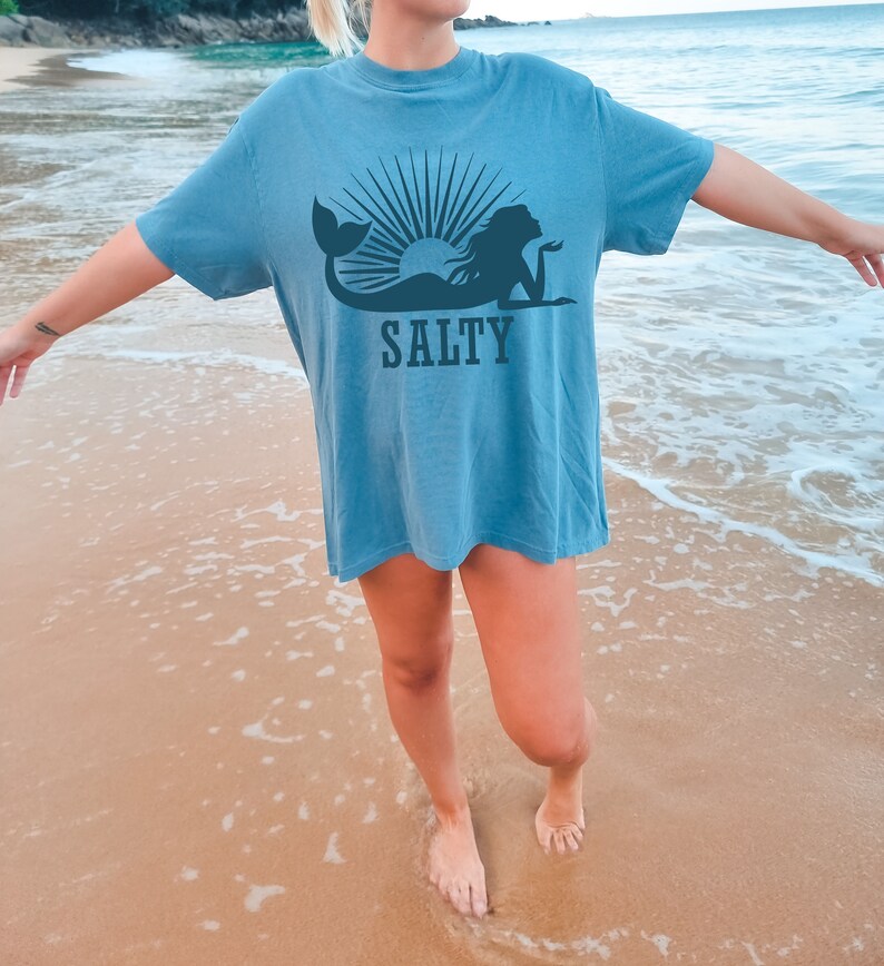 May include: A woman wearing a blue t-shirt with a mermaid and sun graphic and the word "SALTY" printed on it. She is standing on a sandy beach with the ocean in the background.