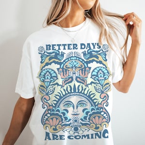 May include: White t-shirt with a colorful graphic design featuring a sun face, butterfly, and floral elements. The text "Better Days Are Coming" is printed on the shirt.