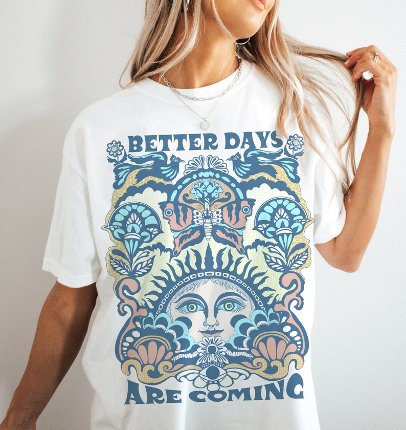 May include: White t-shirt with a colorful graphic design featuring a sun face, butterfly, and floral elements. The text "Better Days Are Coming" is printed on the shirt.