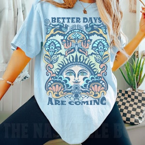 May include: A light blue t-shirt with a colorful graphic design featuring a sun face with a butterfly and the text "Better Days Are Coming".