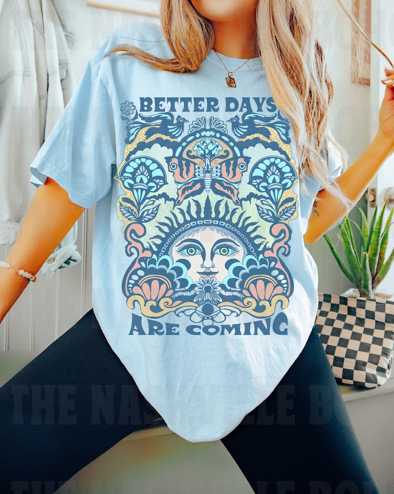 May include: A light blue t-shirt with a colorful graphic design featuring a sun face with a butterfly and the text "Better Days Are Coming".