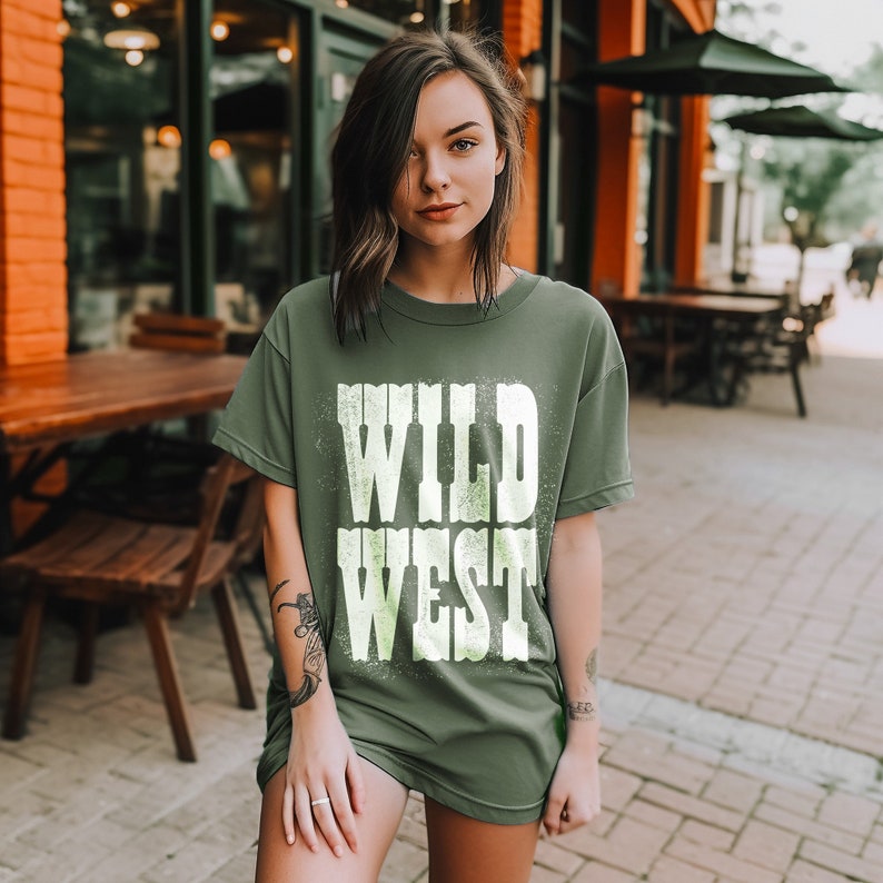Puede incluir: Una camiseta verde con un gr&aacute;fico blanco desgastado que dice "WILD WEST".