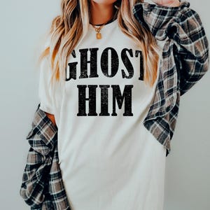 May include: Cream-colored t-shirt with the text "GHOST HIM" in bold, black letters. The person is wearing a black and brown plaid flannel shirt over the t-shirt. The shirt is a casual, everyday wear item.