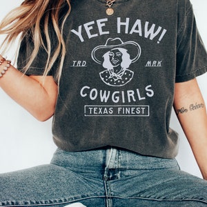 K&ouml;nnte beinhalten: Ein dunkelgraues T-Shirt mit einem wei&szlig;en Grafikdesign, das eine Frau mit Cowboyhut und dem Text "Yee Haw! Cowgirls Texas Finest" zeigt.