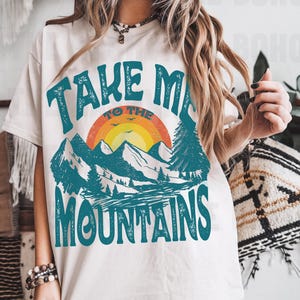 Puede incluir: Camiseta blanca con un gráfico de estilo vintage que dice "Take Me To The Mountains". El gráfico presenta una puesta de sol, montañas y árboles en tonos de azul, verde, naranja y amarillo.