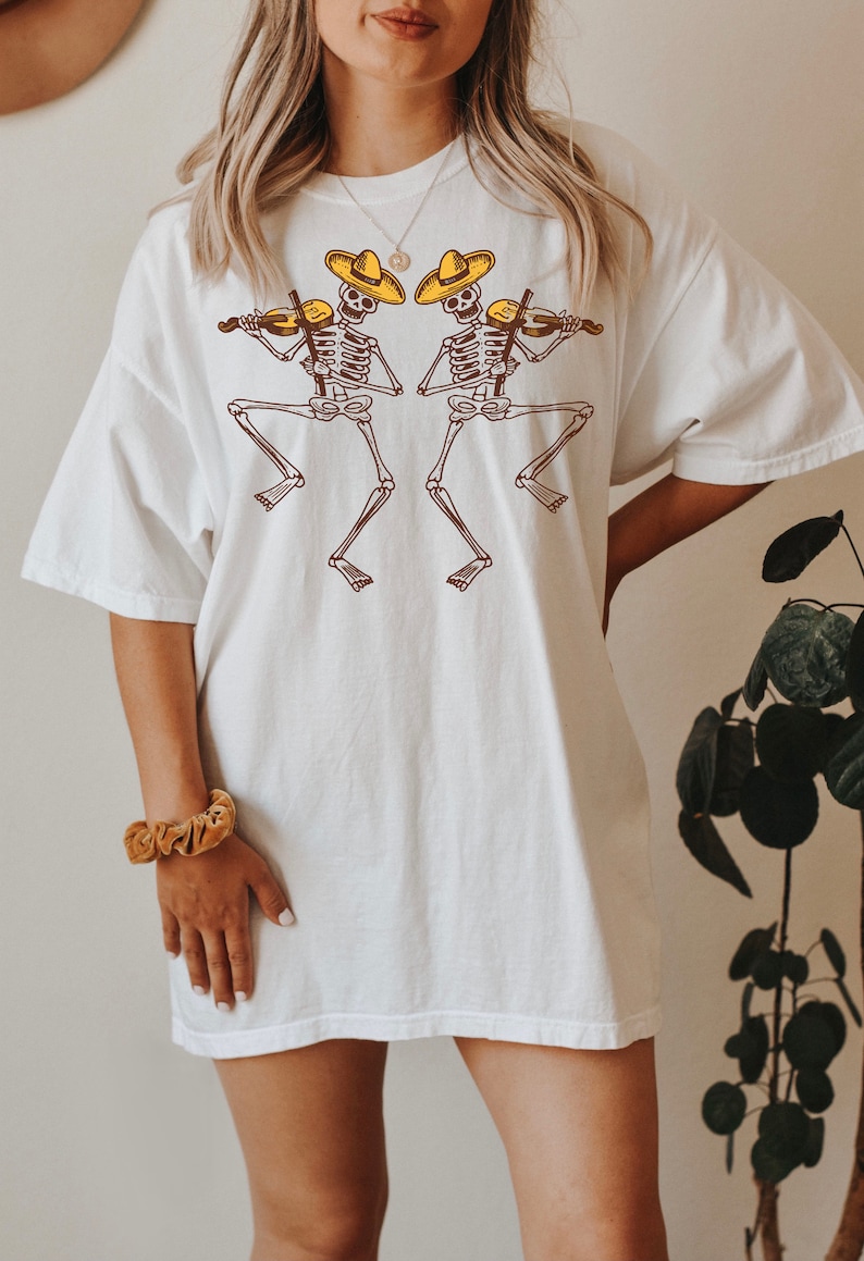 May include: White oversized t-shirt with a graphic of two skeletons wearing sombreros and playing violins.