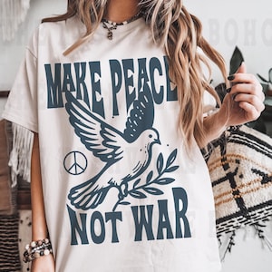 May include: Cream-colored t-shirt with the words "MAKE PEACE NOT WAR" in dark blue, along with a dove carrying an olive branch and a peace symbol. The shirt has short sleeves and a relaxed fit.