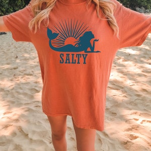 May include: A woman wearing a burnt orange t-shirt with a blue mermaid silhouette and the word "SALTY" printed on it. The woman is standing on a sandy beach.