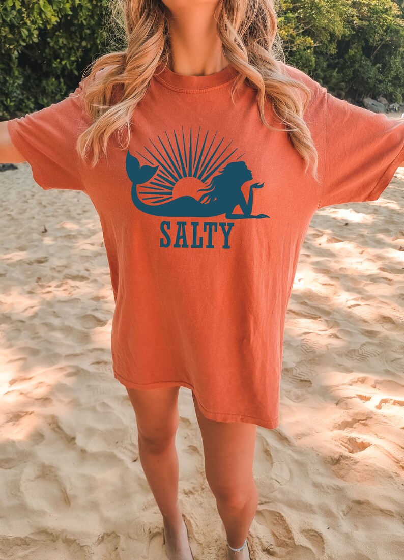 May include: A woman wearing a burnt orange t-shirt with a blue mermaid silhouette and the word "SALTY" printed on it. The woman is standing on a sandy beach.