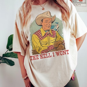 Pode incluir: Uma camiseta de cor creme com uma ilustração vintage de um cowboy usando um chapéu e uma camisa xadrez. O texto "THE HELL I WONT" é impresso abaixo da ilustração.