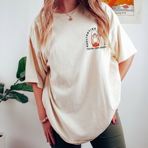 Puede incluir: Una camiseta de color crema con un gr&aacute;fico de una linterna y el texto "Gaslighting You're Just Crazy".
