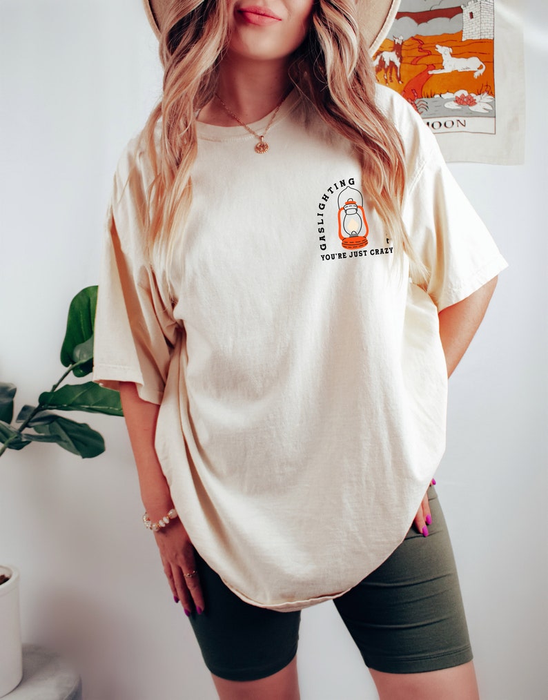 Puede incluir: Una camiseta de color crema con un gr&aacute;fico de una linterna y el texto "Gaslighting You're Just Crazy".