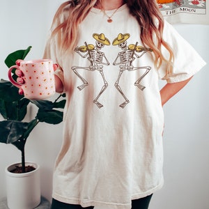 May include: A white oversized t-shirt with a graphic of two skeletons wearing sombreros and playing violins. The skeletons are facing each other and are in a dancing pose.