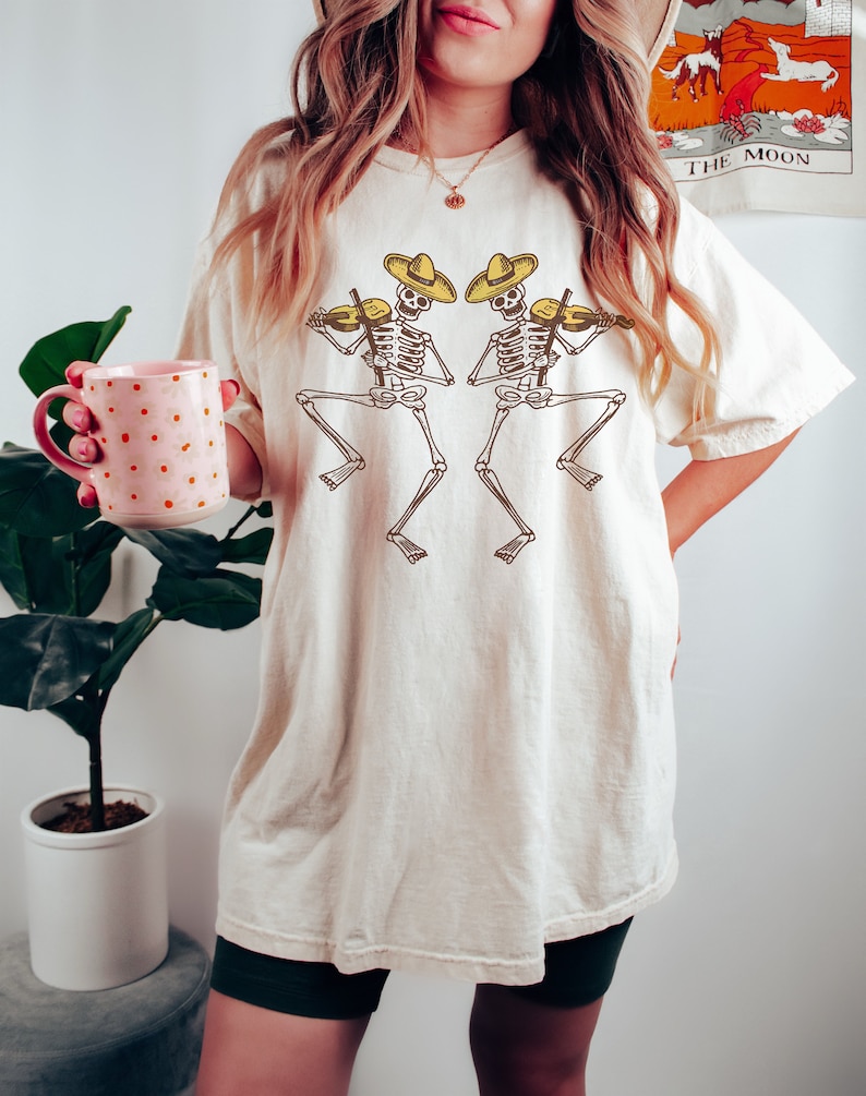 May include: A white oversized t-shirt with a graphic of two skeletons wearing sombreros and playing violins. The skeletons are facing each other and are in a dancing pose.
