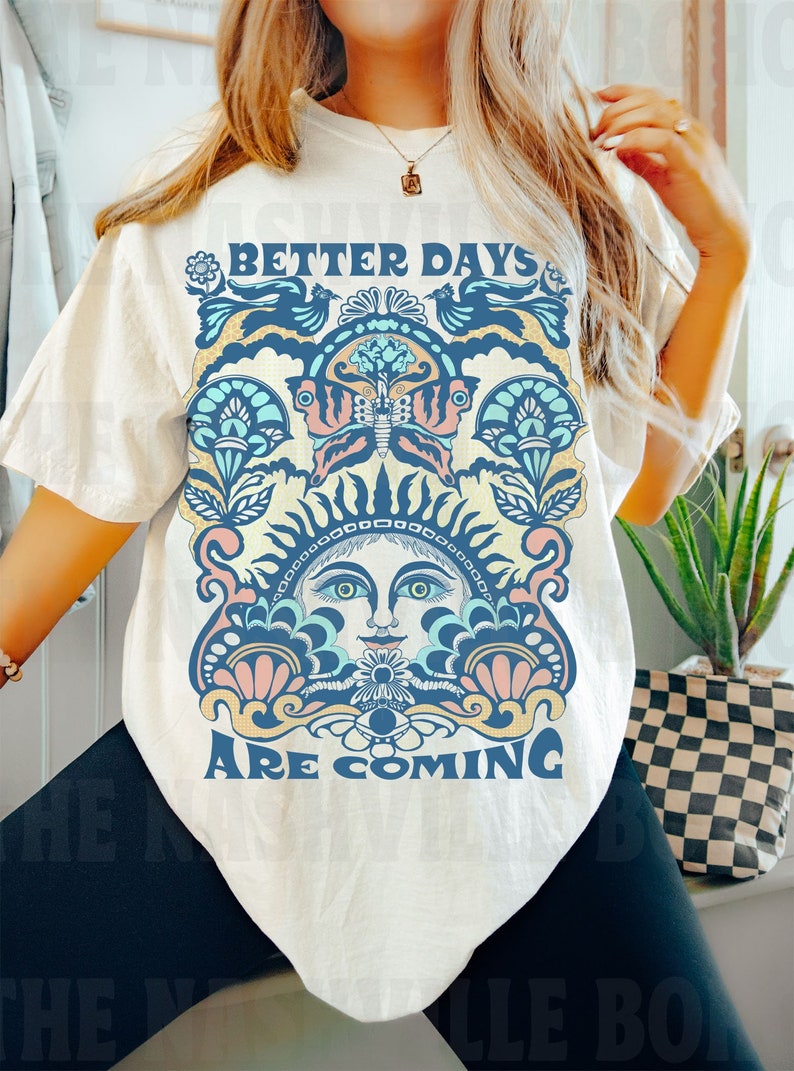 May include: A white t-shirt with a vintage-style graphic design featuring a sun face, butterfly, and floral elements. The text "Better Days Are Coming" is printed on the shirt.