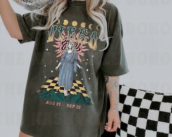 Puede incluir: Una camiseta de manga corta gris carbón con un estampado gráfico del signo zodiacal Virgo. El diseño incluye la palabra VIRGO, fases lunares, una figura con vestido, un patrón de suelo a cuadros, montañas y el texto AUG 23 - SEP 22.