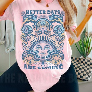 May include: Pink t-shirt with a graphic design featuring a stylized sun with a face and the text "Better Days Are Coming".