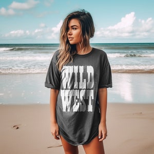 Puede incluir: Una mujer con una camiseta gris oscuro con las palabras "WILD WEST" impresas en blanco en la parte delantera. Est&aacute; de pie en una playa de arena con el oc&eacute;ano al fondo.