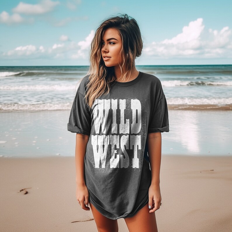 Puede incluir: Una mujer con una camiseta gris oscuro con las palabras "WILD WEST" impresas en blanco en la parte delantera. Est&aacute; de pie en una playa de arena con el oc&eacute;ano al fondo.