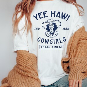 K&ouml;nnte beinhalten: Wei&szlig;es T-Shirt mit einem blauen Grafikdesign, auf dem "Yee Haw! Cowgirls Texas Finest" steht, und einer Zeichnung einer Frau mit Cowboyhut.