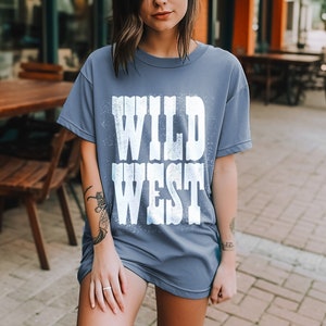 Puede incluir: Una camiseta azul con un gr&aacute;fico blanco que dice "WILD WEST" en una fuente desgastada.