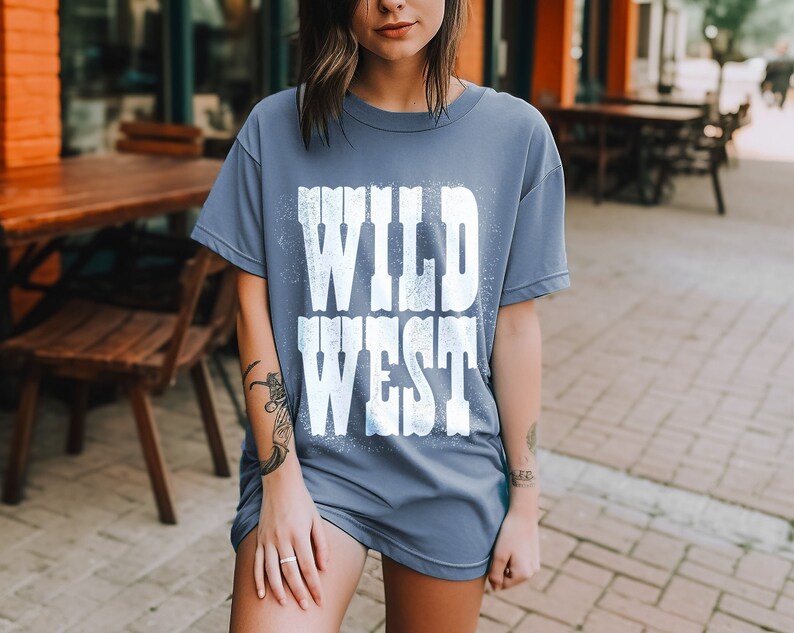 Puede incluir: Una camiseta azul con un gr&aacute;fico blanco que dice "WILD WEST" en una fuente desgastada.