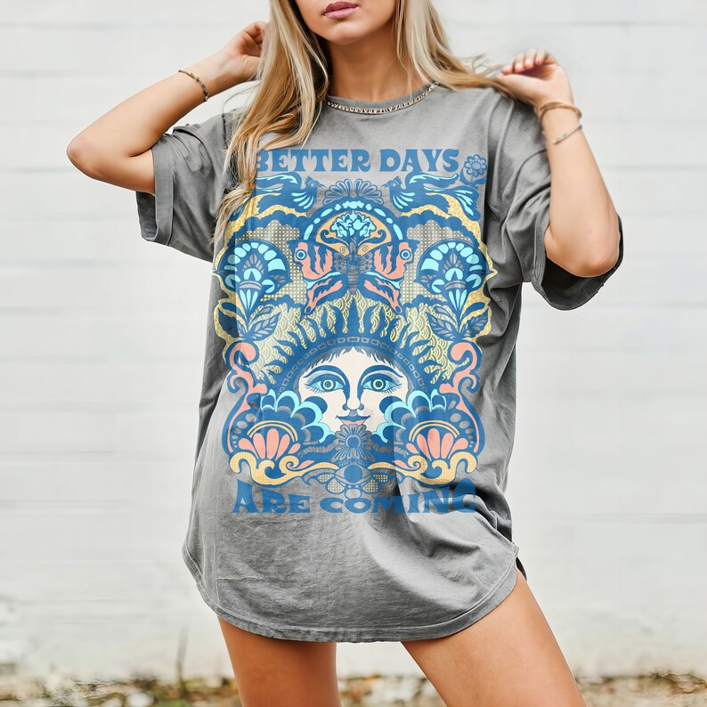 May include: A gray oversized t-shirt with a vintage-inspired graphic design featuring a stylized face surrounded by floral patterns. The text "Better Days Are Coming" is printed on the shirt.