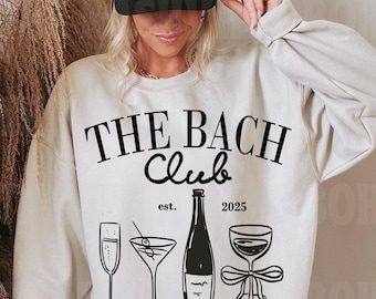 Può includere: Una felpa bianca con testo nero che recita "THE BACH Club est. 2025" e una grafica di quattro bevande: un flute di champagne, un bicchiere da martini, una bottiglia di vino e un bicchiere di vino con un fiocco. Sotto la grafica, il testo "Lauren's Bachelorette Nashville, TN".