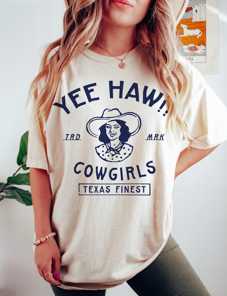 K&ouml;nnte beinhalten: Ein wei&szlig;es T-Shirt mit einer blauen Grafik einer Frau mit Cowboyhut und dem Text "Yee Haw!" "TRD" "MARK" "COWGIRLS" "TEXAS FINEST"