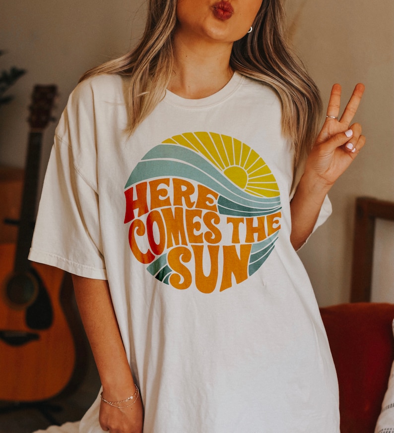 K&ouml;nnte beinhalten: Wei&szlig;es T-Shirt mit einem Retro-Grafikdesign einer Sonne, Wellen und dem Text "Here Comes The Sun".