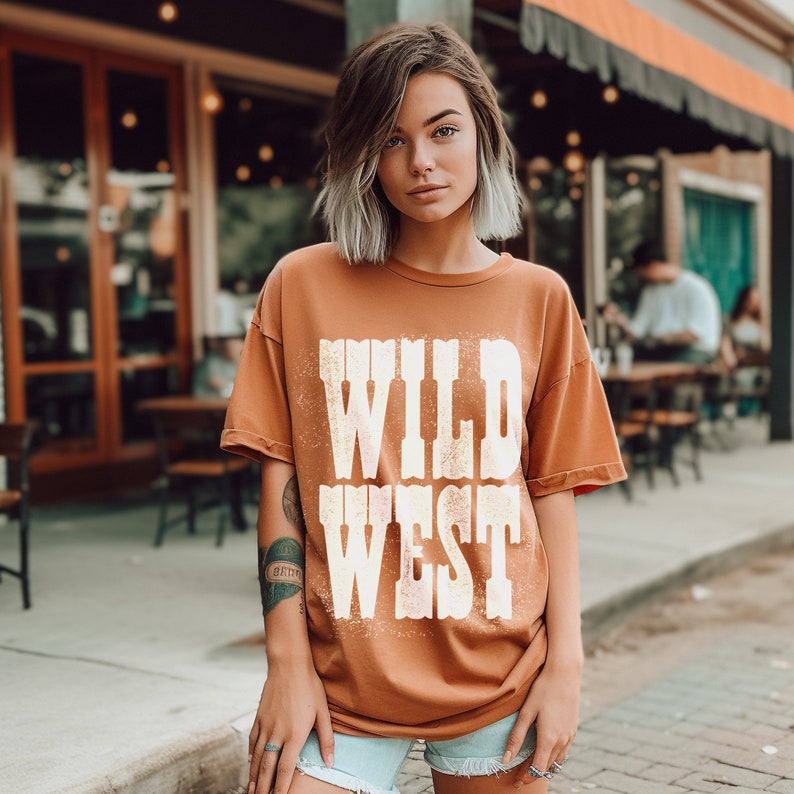 Puede incluir: Una mujer lleva una camiseta marr&oacute;n con el texto "WILD WEST" impreso en blanco. Est&aacute; de pie frente a un edificio con una fachada de ladrillo.