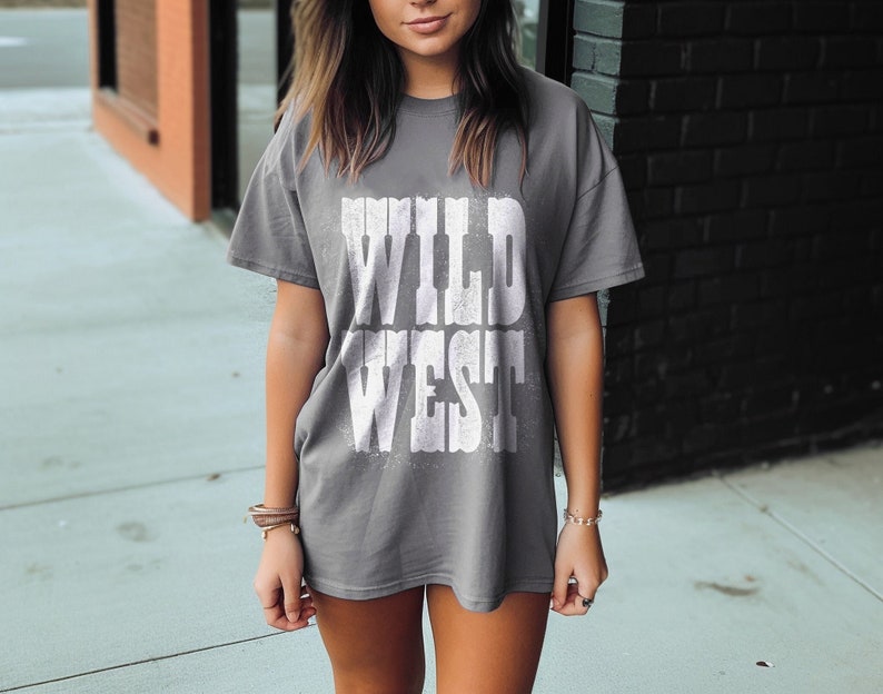 Puede incluir: Una camiseta gris con las palabras "WILD WEST" impresas en blanco en la parte delantera.