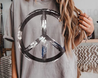 May include: A grey short-sleeve t-shirt featuring a large, distressed black and white peace sign graphic centered on the chest. The design has a textured, brushstroke appearance with visible white highlights against the dark grey fabric.