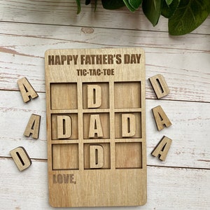 May include: A wooden tic-tac-toe game with the word "DAD" in the center. The game is designed for Father's Day and includes the words "Happy Father's Day" and "Tic-Tac-Toe" at the top. The game also includes the words "Love," and comes with wooden letter tiles.