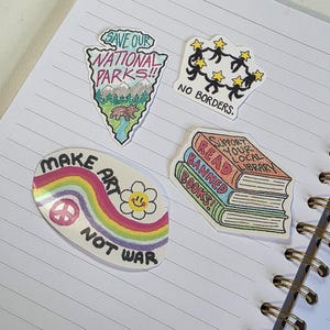 May include: Four vinyl stickers featuring various designs: a shield-shaped sticker with a bear and the text SAVE OUR NATIONAL PARKS, a circular sticker with a rainbow and the text MAKE ART NOT WAR, a stack of books with the text READ BANNED BOOKS, and a circular sticker with figures and the text NO BORDERS.