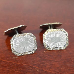 May include: A pair of vintage silver cufflinks with an octagonal shape. The cufflinks have a detailed engraved design on the face and a silver finish. The cufflinks are displayed on a dark brown wooden surface.