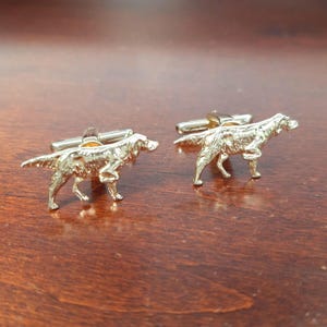 May include: A pair of gold-colored cufflinks, each shaped like a dog in a walking pose. The cufflinks have a shiny finish and are set against a dark wood surface. The dog figures are detailed, showing the breed's features.