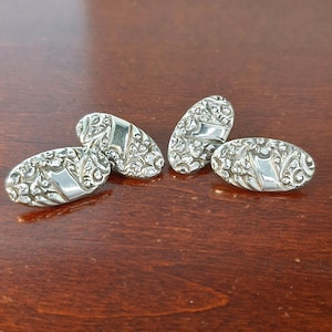 May include: Four silver oval cufflinks with ornate floral and scrollwork designs. Each cufflink has a rectangular center and a curved, textured surface. The cufflinks are displayed on a dark brown wooden surface.